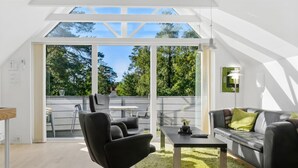 Una sala moderna con un sillón de cuero negro, un sofá gris y una mesa de centro. Hay un gran ventanal que da a un patio exterior.