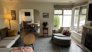 Sala de estar con sofá, reposapiés, mesa de centro y chimenea. Hay un comedor con mesa y sillas, y una cocina al fondo.