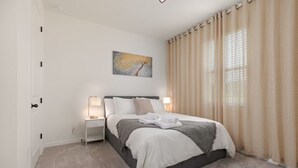 A neatly arranged bedroom with a bed, bedside lamps, a painting, and a window with curtains.