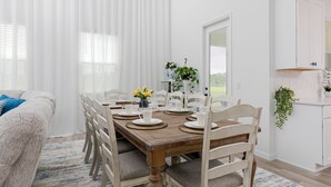 A dining area with a wooden table set for six, surrounded by white chairs with grey cushions.