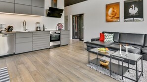 Une cuisine moderne avec des meubles gris, un four encastré et un téléviseur à écran plat. On y trouve un canapé en cuir noir avec un coussin rouge et une table basse en verre, sur laquelle reposent un bol et une télécommande.