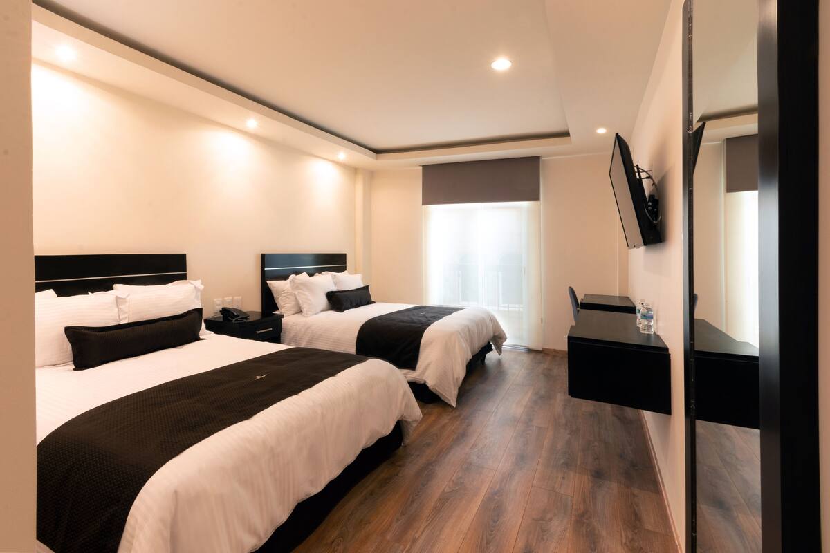 A hotel room with two beds, a bedside table with a phone, a small table with a glass of water, and a mounted TV.