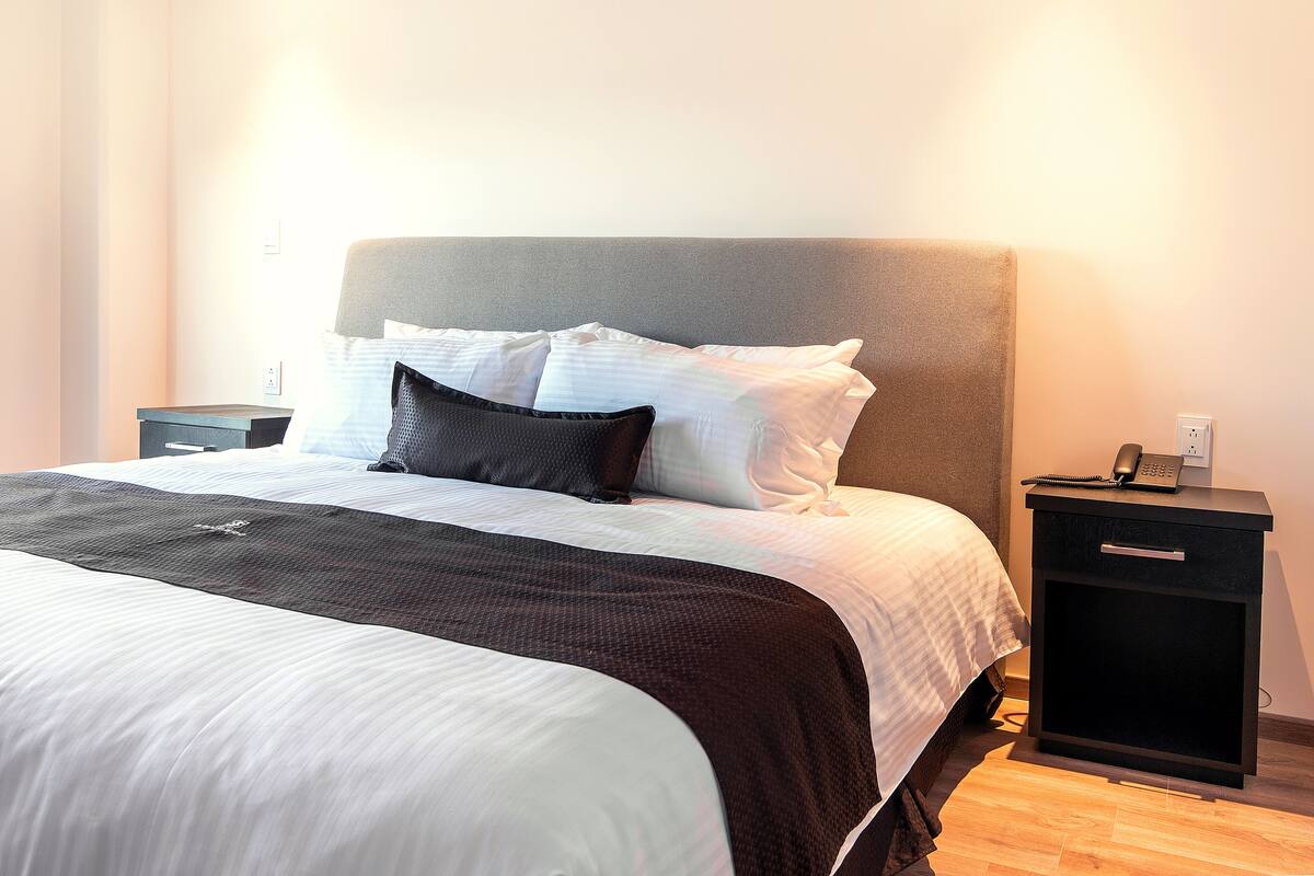 A neatly made bed with white and dark bedding, a bedside table with a phone, and a wall socket.