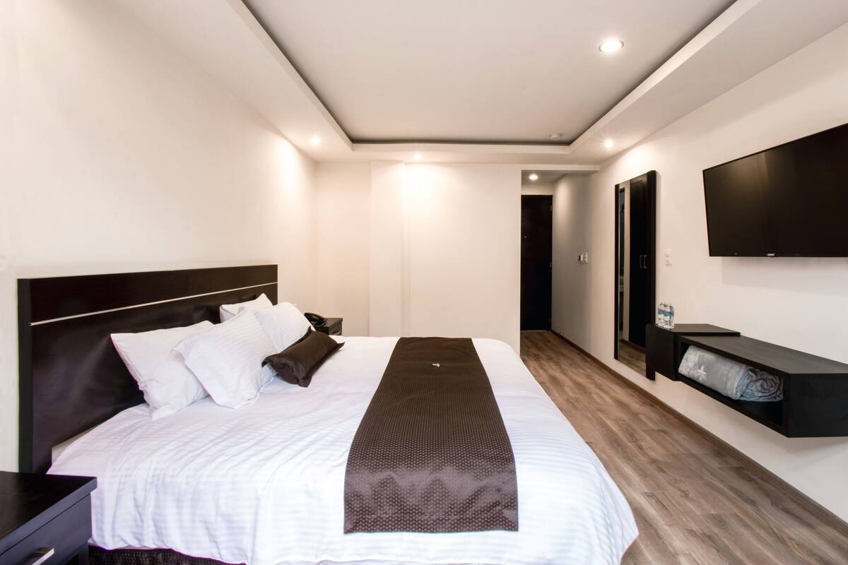 A modern hotel room with a large bed, a flat-screen TV mounted on the wall, and a small shelf with a bottle.