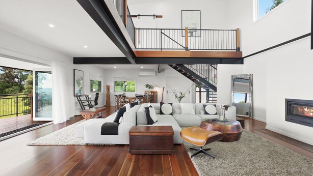 Un salon spacieux avec un grand canapé d’angle, une table basse en bois et une cheminée. Un escalier mène à un niveau supérieur qui donne sur un balcon.