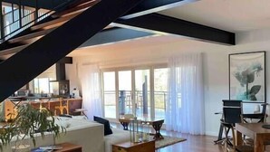 Un vaste espace de vie agrémenté d’un escalier en bois, d’un coin repas et d’une cuisine visible à travers de grandes fenêtres.