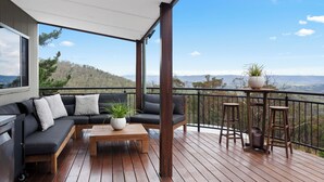 Une terrasse couverte avec un canapé de couleur foncée, une table en bois et deux tabourets de bar.