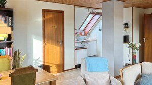 Een gezellige woonkamer met een houten eettafel, twee fauteuils en een bank. Door een open deur is een keuken zichtbaar.