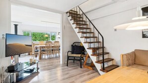 A modern living room with a wooden staircase, a dining area with chairs, and a TV on a stand.