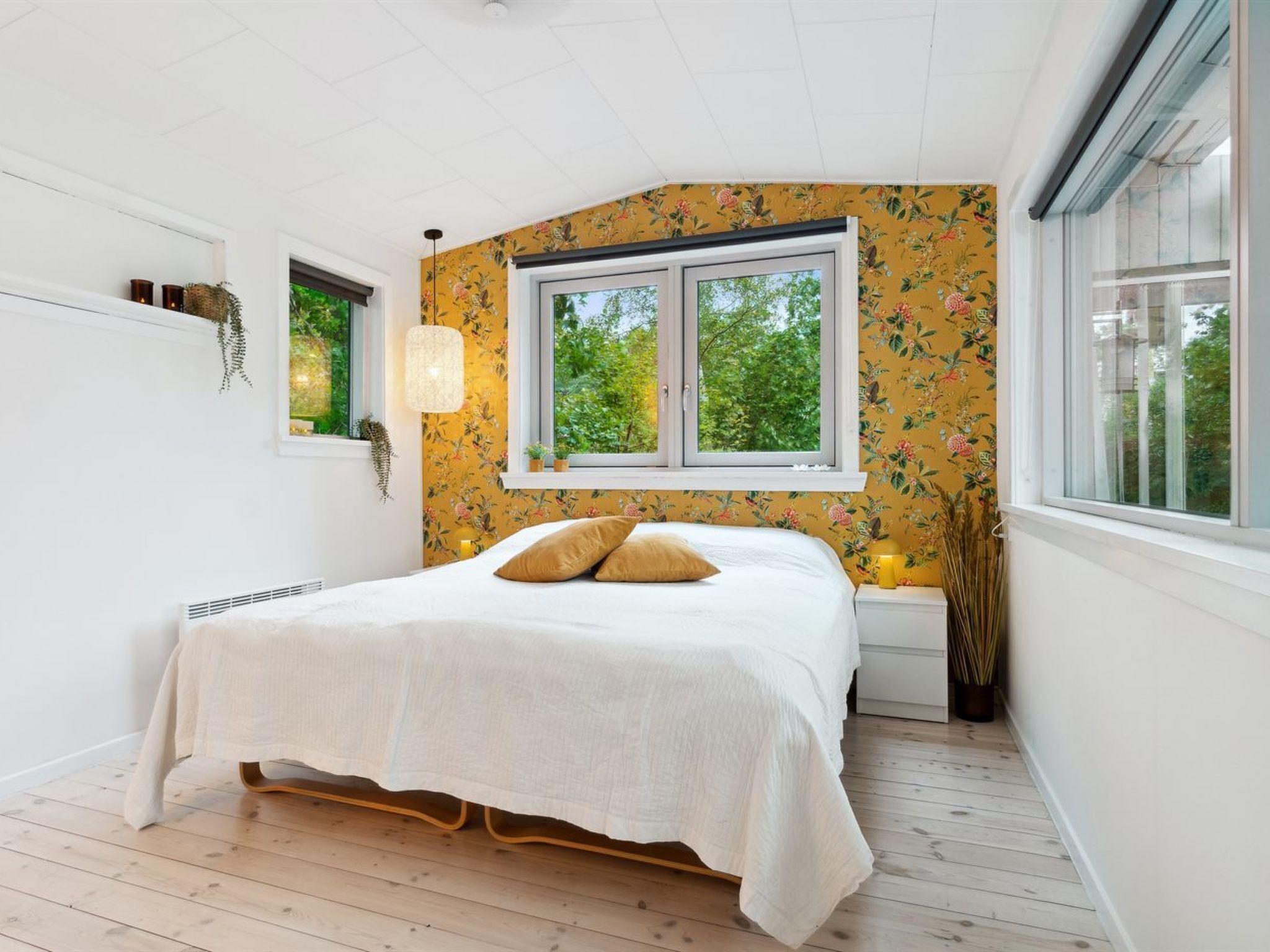 Ett sovrum med en säng, ett fönster, ett element och en vägg med blommig tapet.