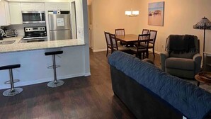 A modern kitchen with white cabinets, stainless steel appliances, and a bar area with stools. There is a dining area with a wooden table and chairs, and a blue sofa in the living room. - Perfect 2/1 beach duplex apartment in the heart of STA beach (St. Augustine)