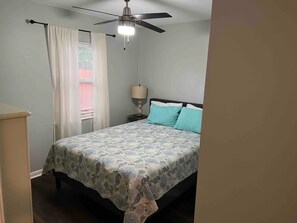 Un dormitorio con cama, ventilador de techo, mesita de noche y ventana con cortinas.