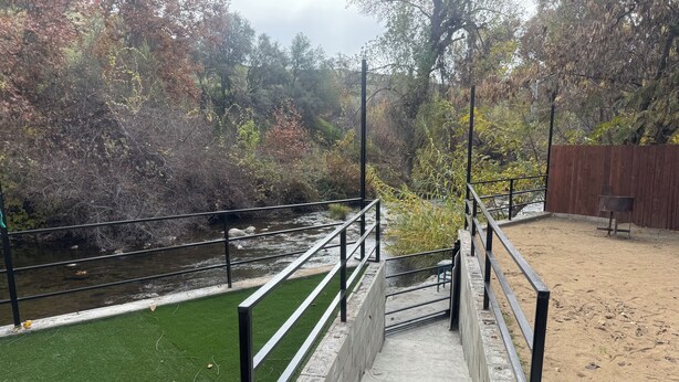 A concrete staircase leading to a sandy area with a metal railing, a small grassy patch, and a river with rocks.