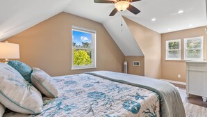 A bedroom with a sloped ceiling, a bed with a floral bedspread, a ceiling fan, and a window with a view of greenery.