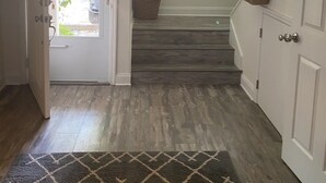 Un pasillo con piso de madera, una alfombra, una puerta que da al exterior, una ventana con persianas y una escalera.
