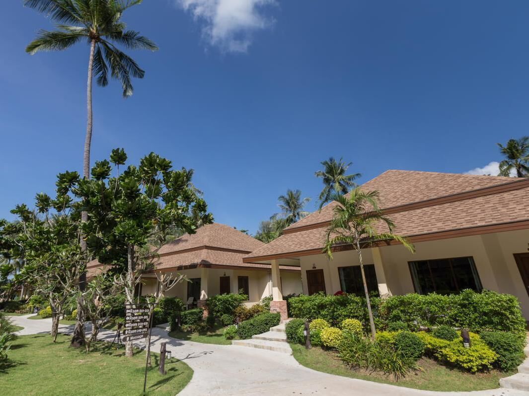 A resort with multiple bungalow-style buildings, a well-maintained garden with palm trees and shrubs, and a clear blue sky.