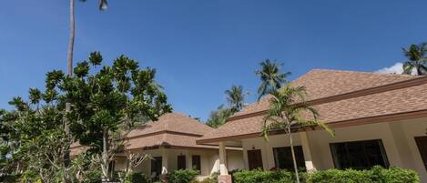 A resort with multiple bungalow-style buildings, a well-maintained garden with palm trees and shrubs, and a clear blue sky.