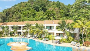 Een resort met een groot zwembad, palmbomen en een heuvellandschap op de achtergrond.