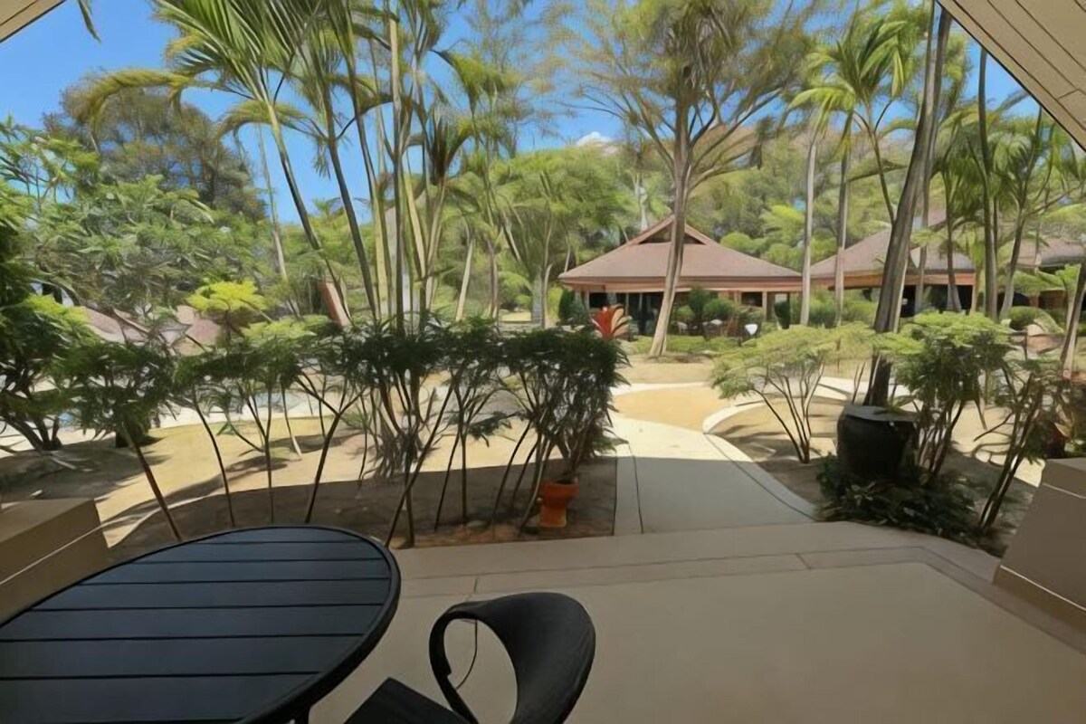 Un patio con una mesa y sillas, rodeado de plantas tropicales y un edificio de techo marrón.