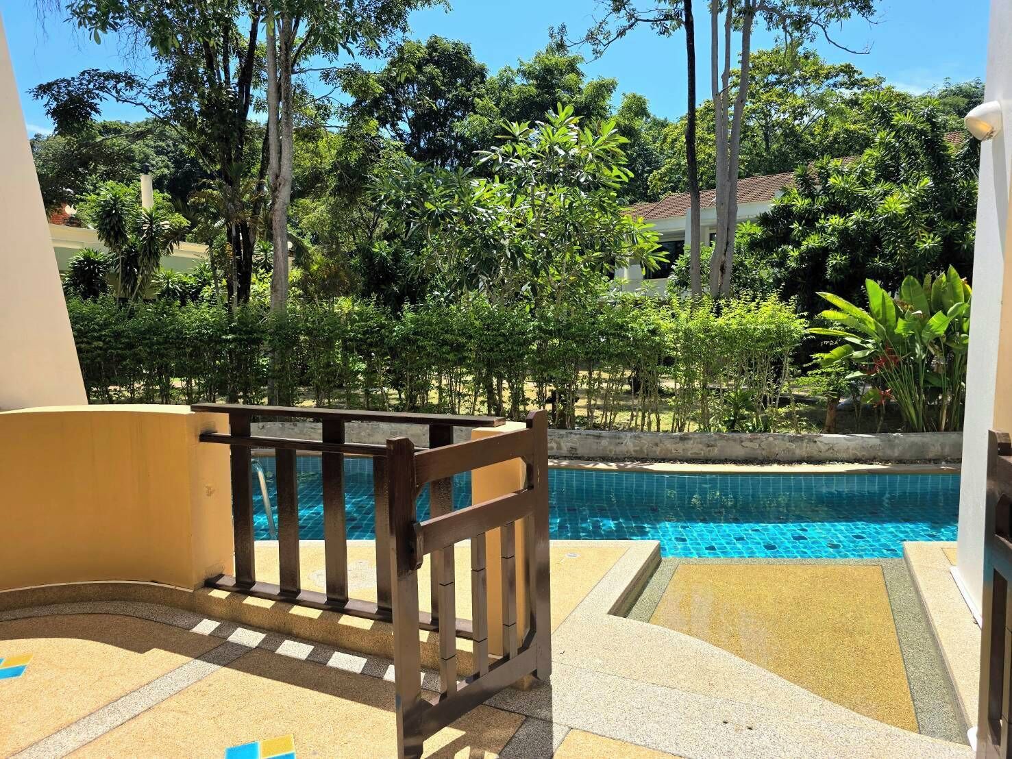 Un espace piscine doté d’une terrasse en bois et offrant une vue sur une végétation luxuriante et des bâtiments en arrière-plan.