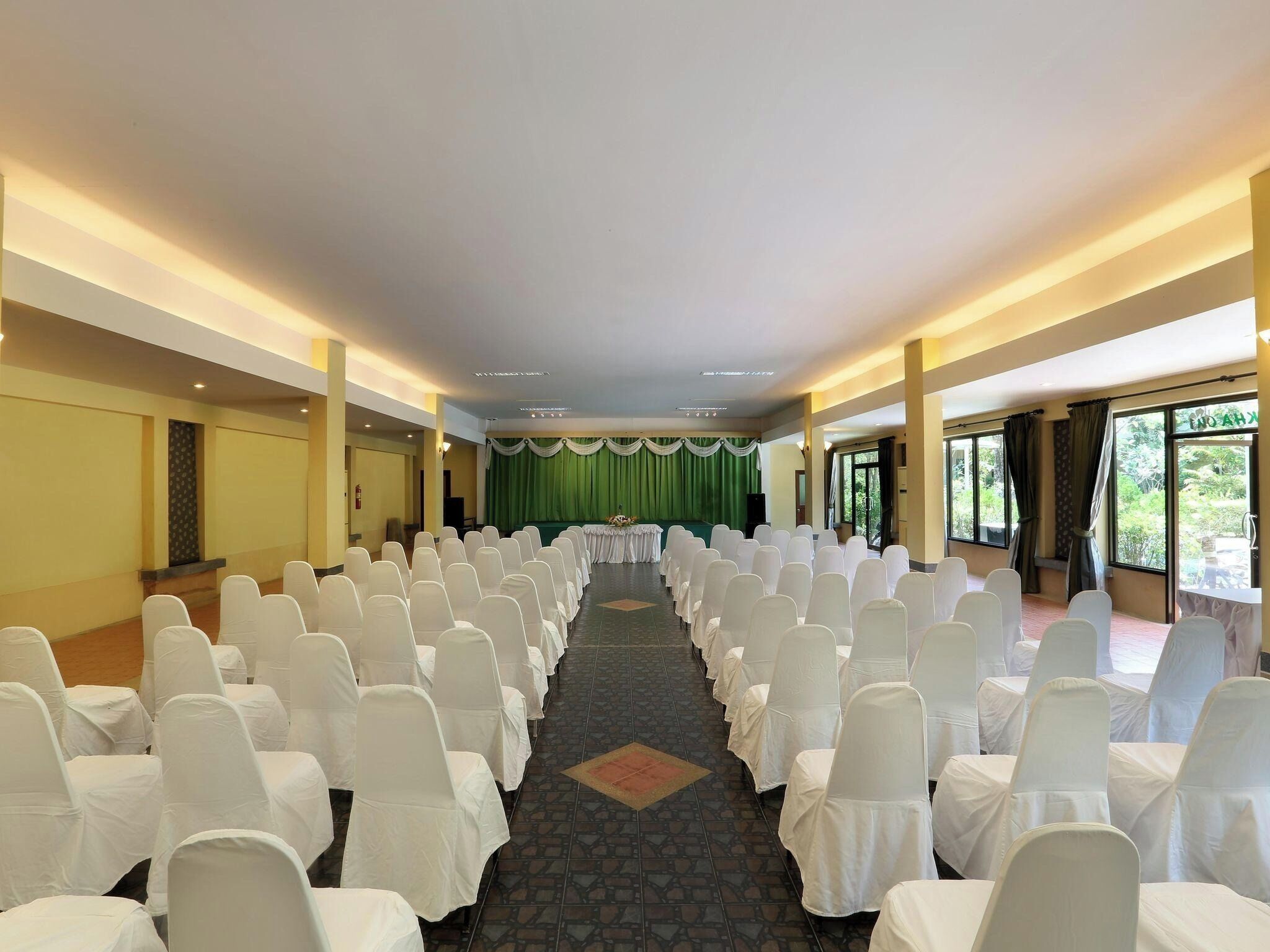 A conference room with rows of white chairs, a stage with a green curtain, and large windows offering a view of greenery.