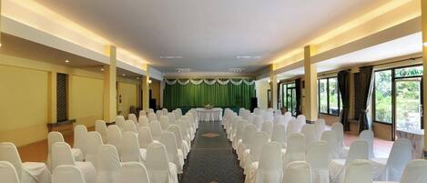 A conference room with rows of white chairs, a stage with a green curtain, and large windows offering a view of greenery.