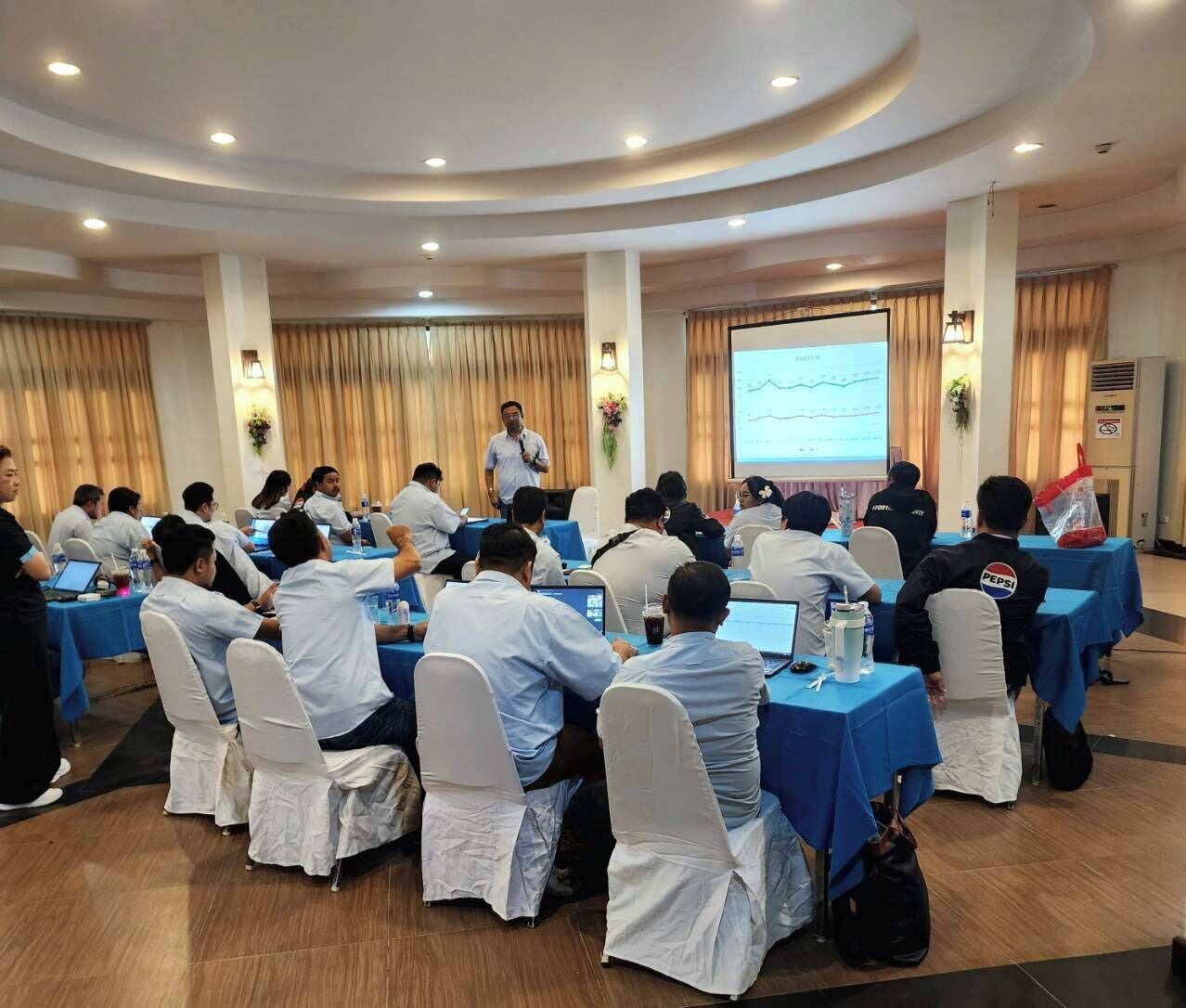 A conference room with round tables, attendees seated, and a presenter at the front.