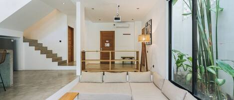 A modern interior with a sofa, wooden bench, and a round table in a well-lit space with a large window and staircase.