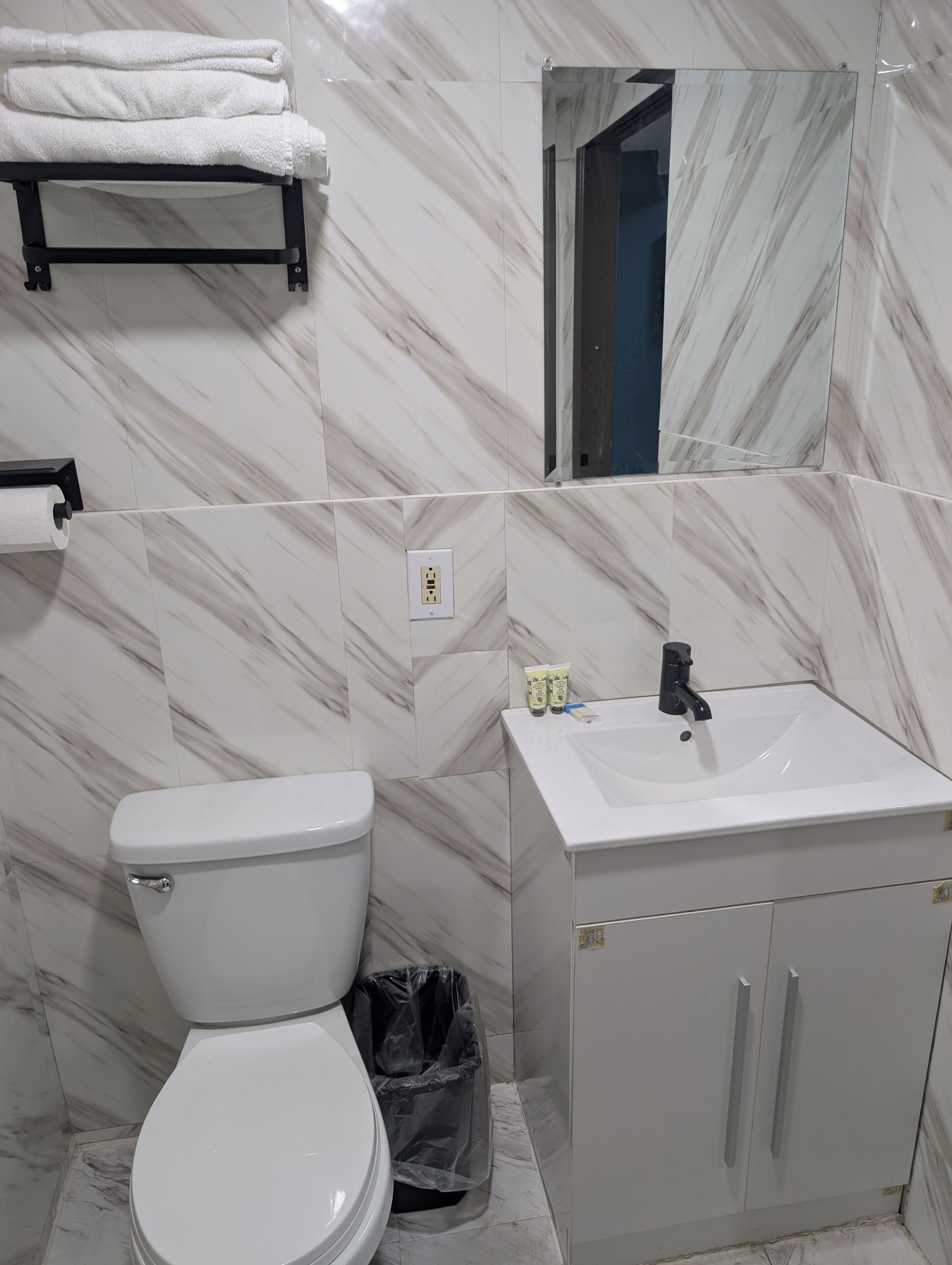 A bathroom with a toilet, sink, mirror, towel rack, and trash bin.