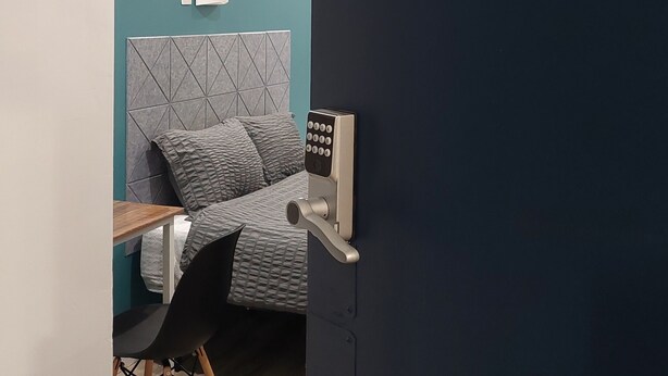 A room with teal walls, a bed with a geometric headboard, a wooden table, a black chair, and a door with a number plate. - APD Hotel (Montreal)