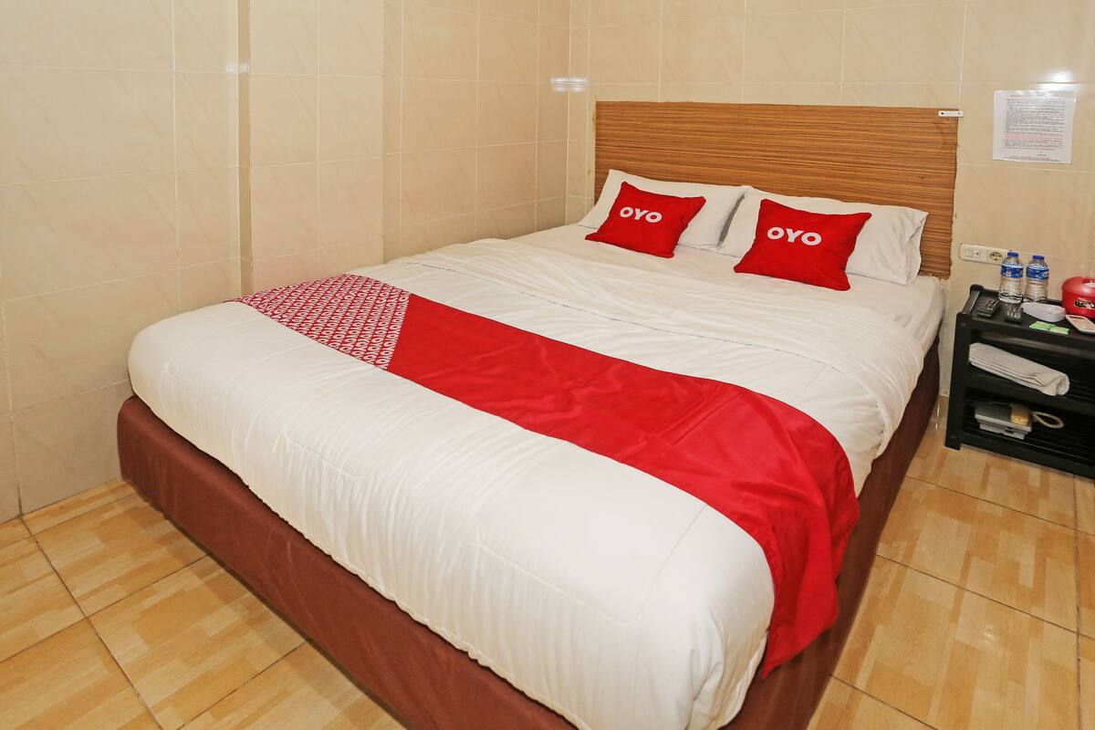 A neatly made bed with white and red bedding, a wooden headboard, and a nightstand with a remote control and water bottles.