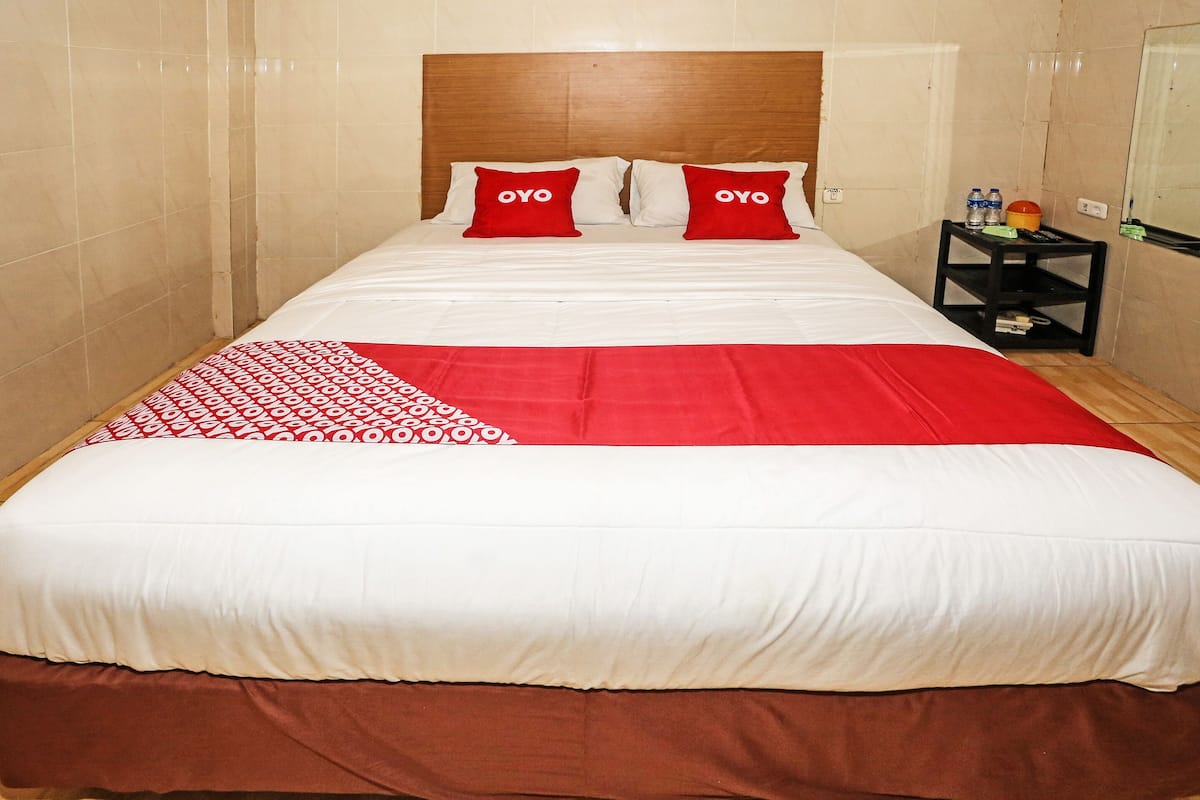A hotel room with a large bed, two red pillows with the word "OYO" on them, a red and white patterned throw, and a small black nightstand with a bottle and a toothbrush.