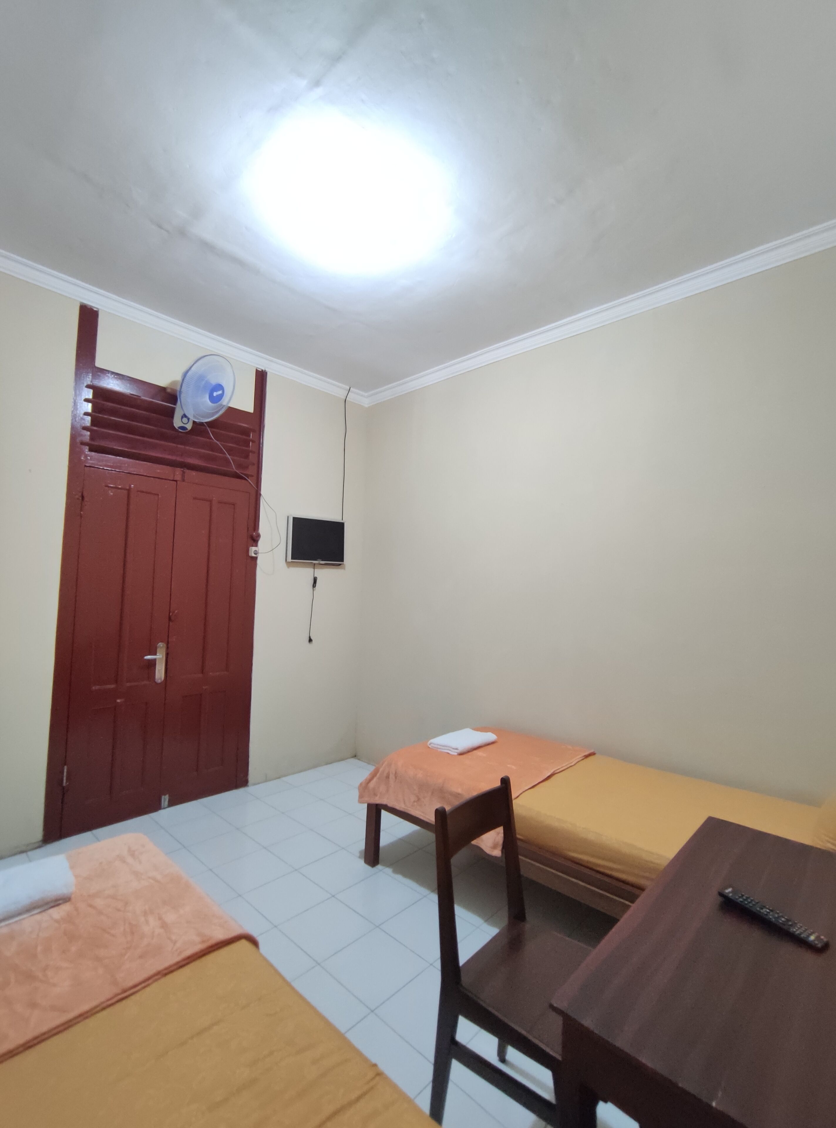 Quarto pequeno e modesto com uma cama, uma cadeira, uma mesa, uma televisão e uma porta.