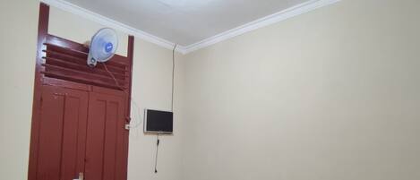 Quarto pequeno e modesto com uma cama, uma cadeira, uma mesa, uma televisão e uma porta.