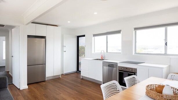 A modern kitchen with white cabinetry, stainless steel appliances, and a dining area with a wooden table and wicker chairs. - Coastal Breeze (Ulladulla)
