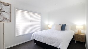 A bedroom with a bed, two bedside tables with lamps, a window with blinds, and a wall-mounted artwork. - Coastal Breeze (Ulladulla)