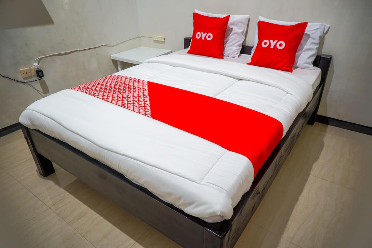 A bed with white and red bedding, two pillows with the word "OYO" on them, a white nightstand, and a wall socket.