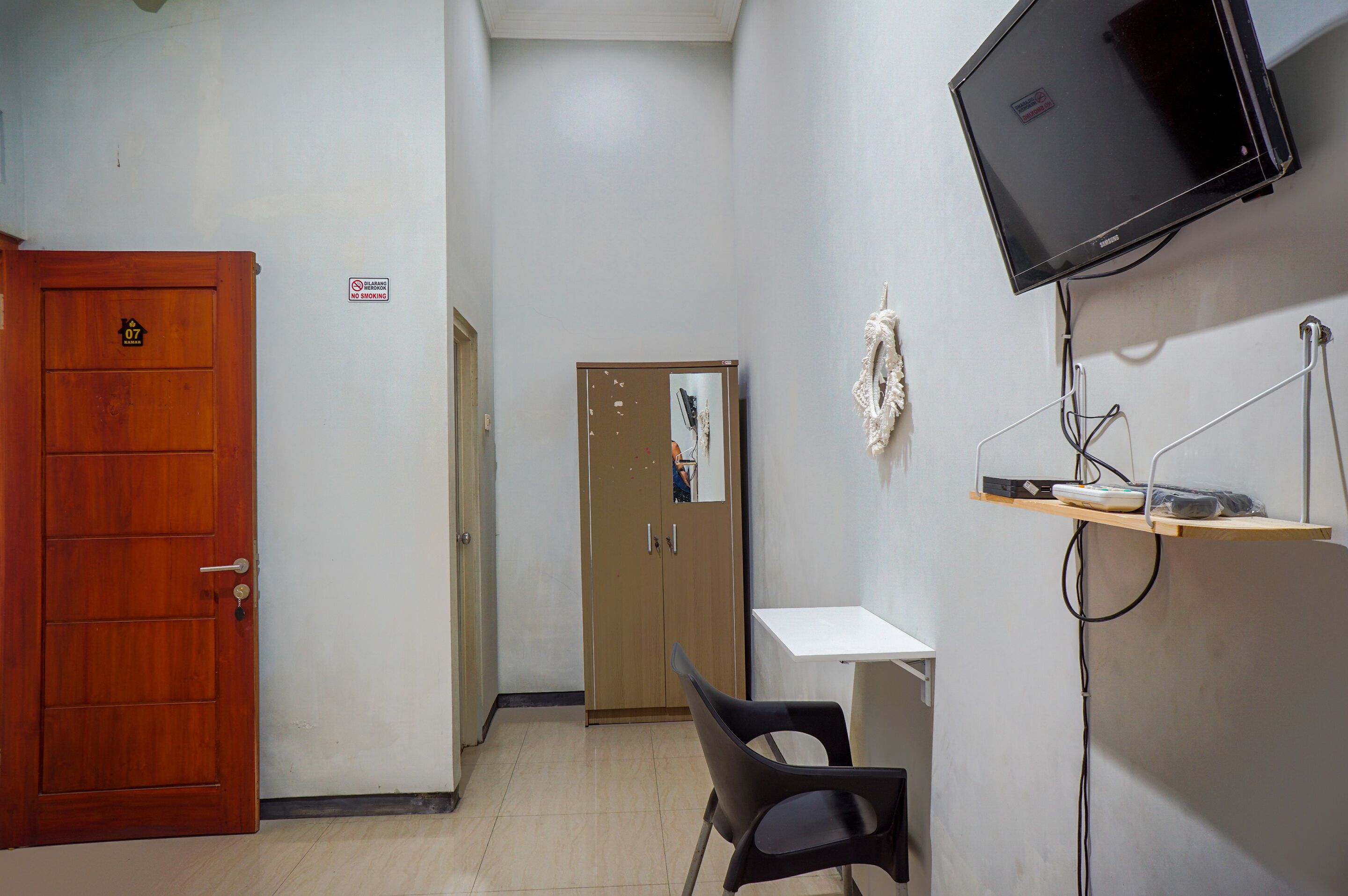 Camera d'albergo con porta in legno, una scrivania bianca, una sedia nera, una televisione a muro e un piccolo ripiano con uno specchio e un asciugamano.