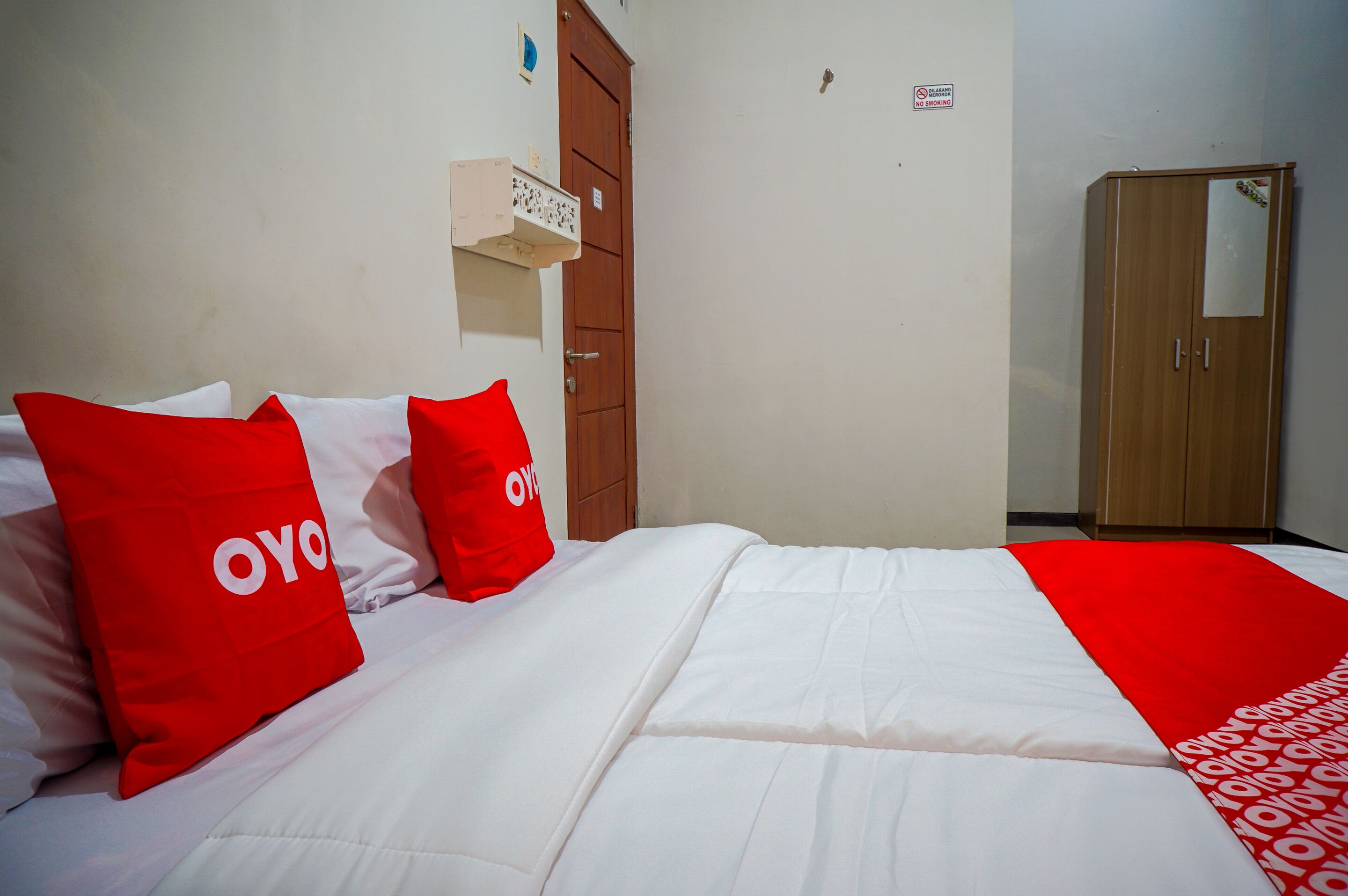 A hotel room with a bed, red pillows, a wooden wardrobe, and a white wall.