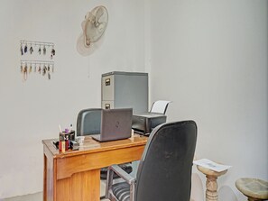 Um escritório pequeno com uma mesa, um laptop, uma cadeira, um armário de arquivos e um ventilador fixado na parede.