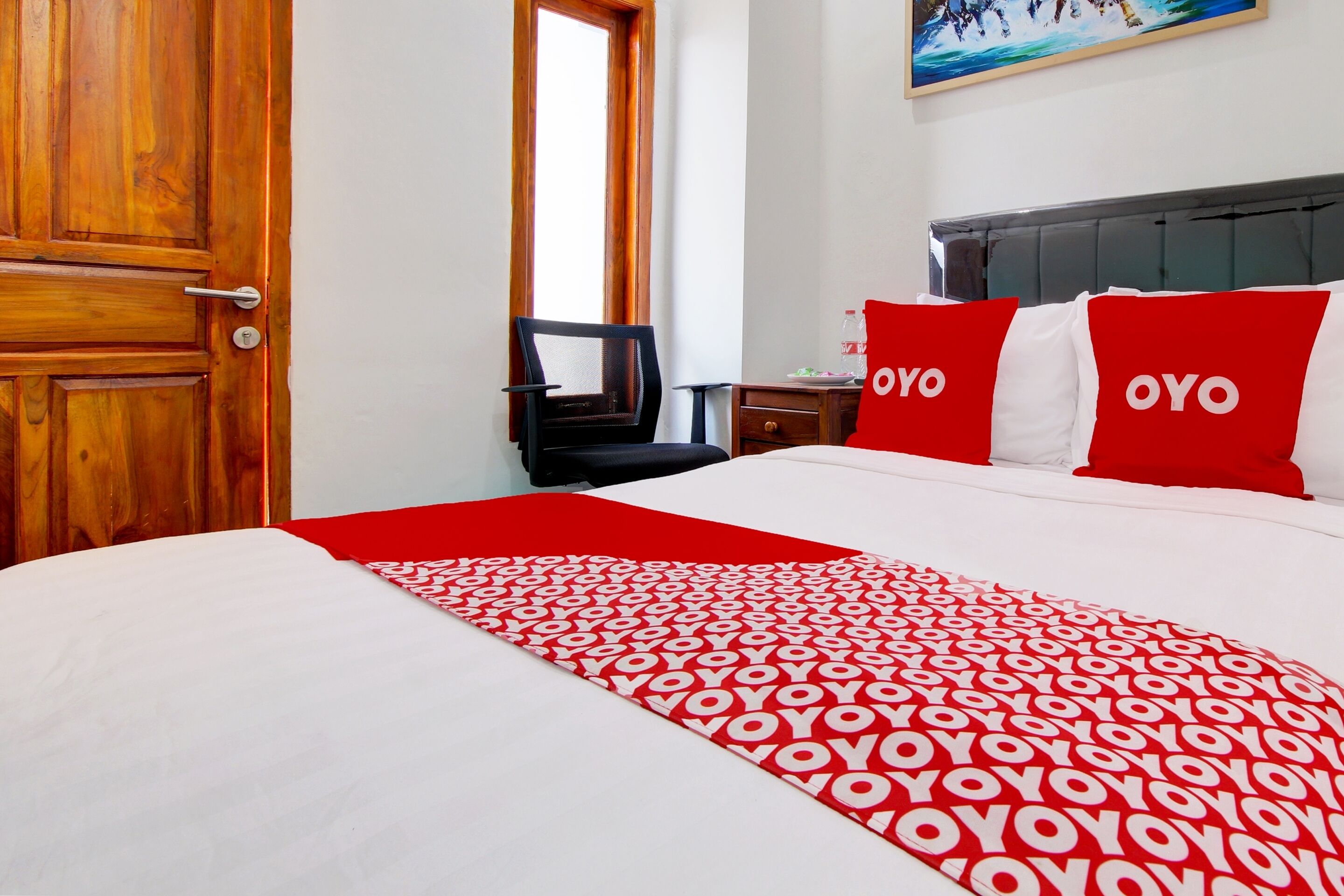 A hotel room with a bed, a wooden door, a chair, a nightstand, and a painting on the wall.