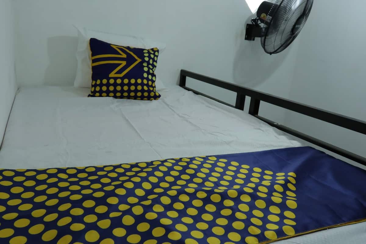 A bed with a white mattress, a blue and yellow polka dot blanket, and a pillow with a yellow arrow design. A wall-mounted fan is visible to the right.