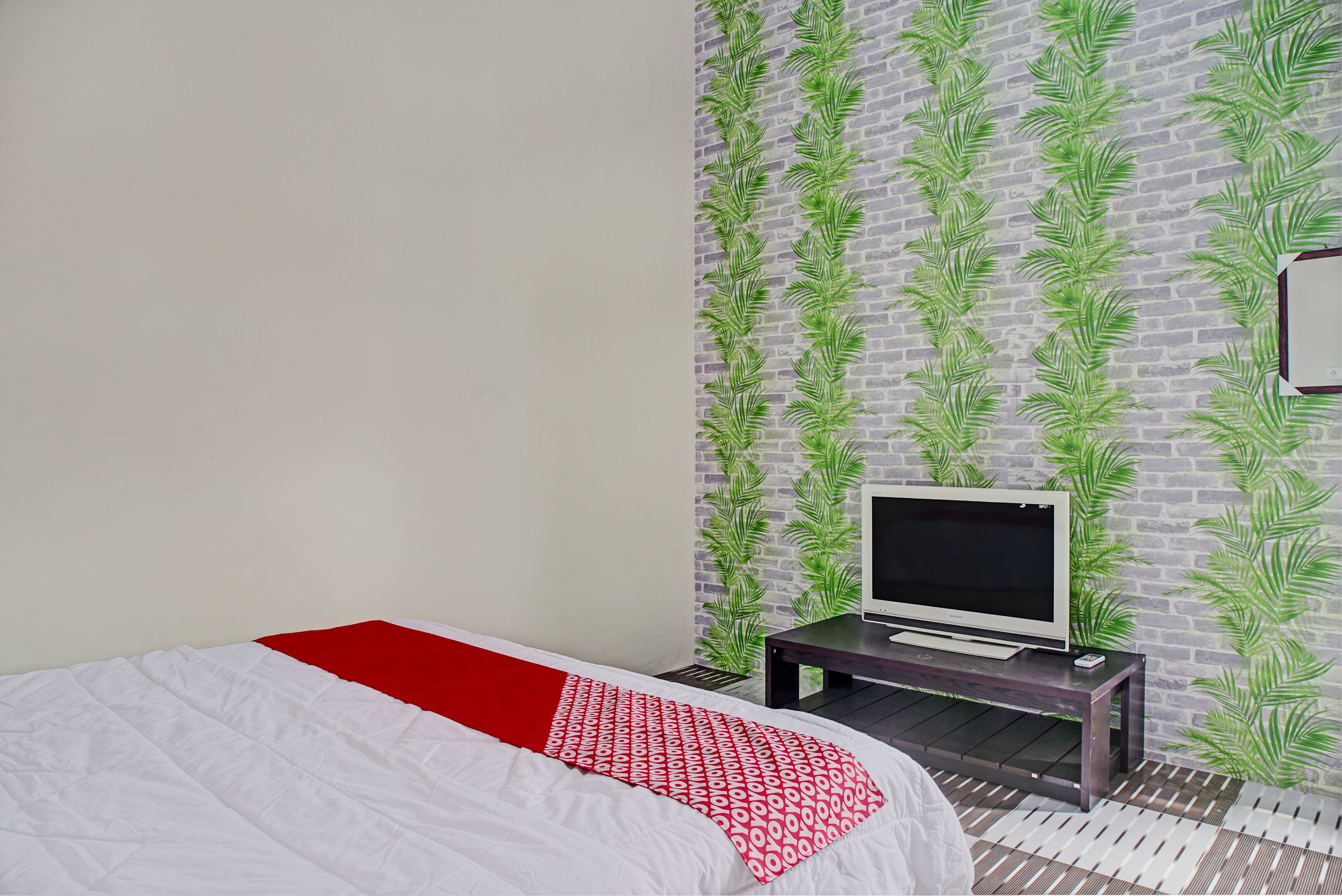 A bedroom with a bed, a TV on a stand, and a brick-patterned wall with green foliage.