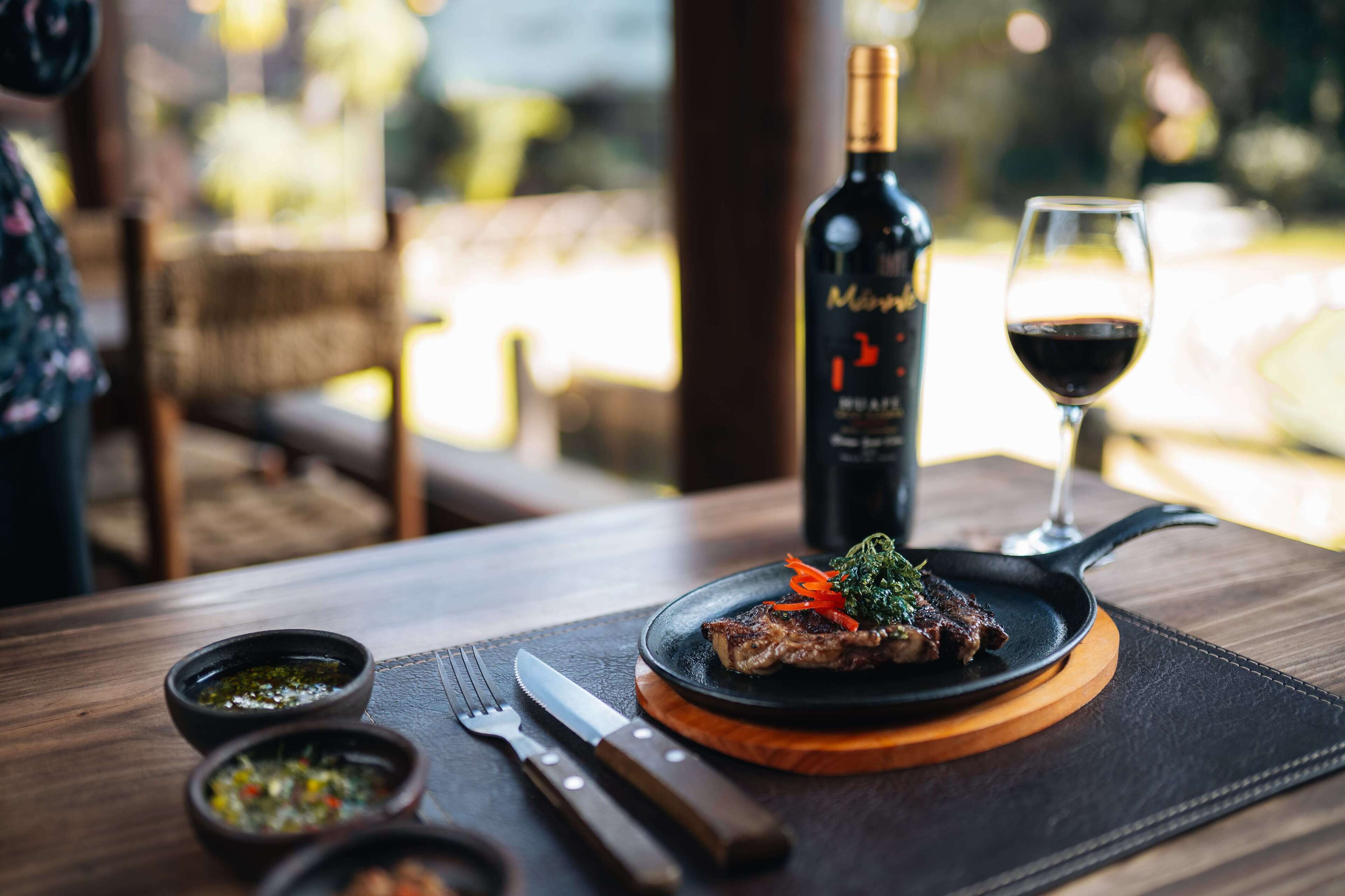A dining table with a steak, a bottle of wine, and a glass of wine.