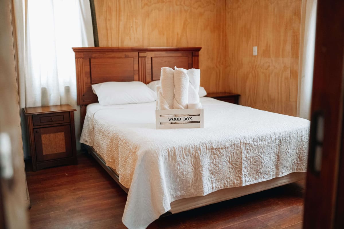Ein ordentlich bezogenes Bett mit weißer Bettdecke und einem hölzernen Kopfteil. Auf dem Bett liegt eine weiße Schachtel mit der Aufschrift 'WOOD BOX'.