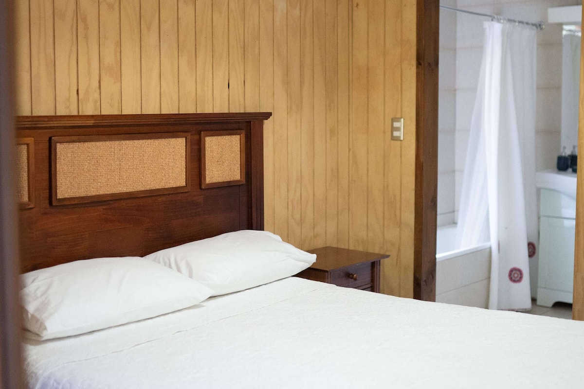 Ein Zimmer mit Holzwänden, einem Bett, einem Nachttisch und einem durchgegangenen Tür angedeuteten Badezimmer.