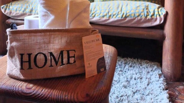 A cozy cabin interior with a wooden bench, a basket labeled "HOME" containing towels, and a window with a view of greenery. - Complejo Turístico Valle del Sol Quillón (Quillón)