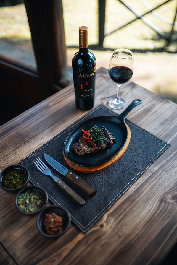 A wooden table with a black plate holding a steak, a bottle of wine, and a glass of red wine. - Complejo Turístico Valle del Sol Quillón (Quillón)
