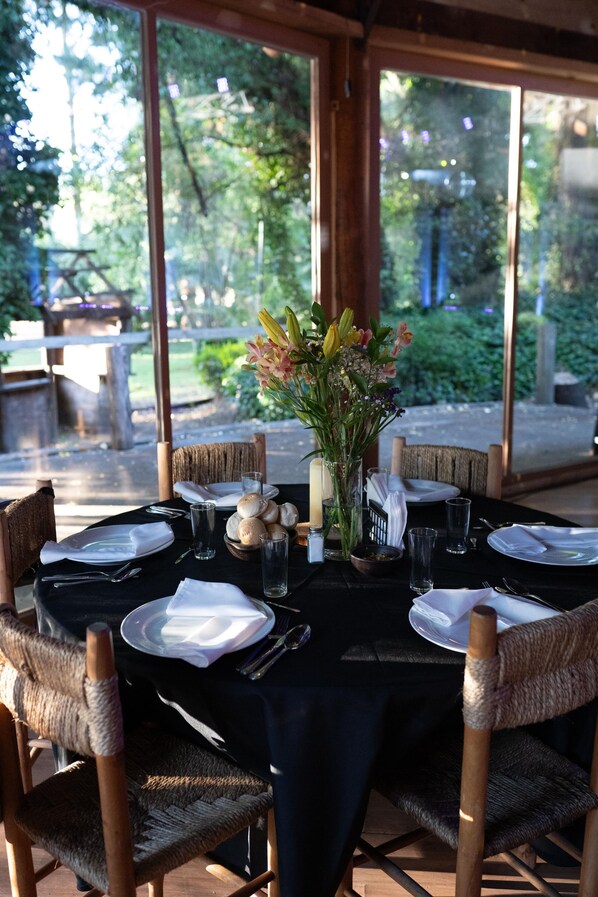 A dining table set for six with wicker chairs, a vase of flowers, and a view of greenery outside. - Complejo Turístico Valle del Sol Quillón (Quillón)