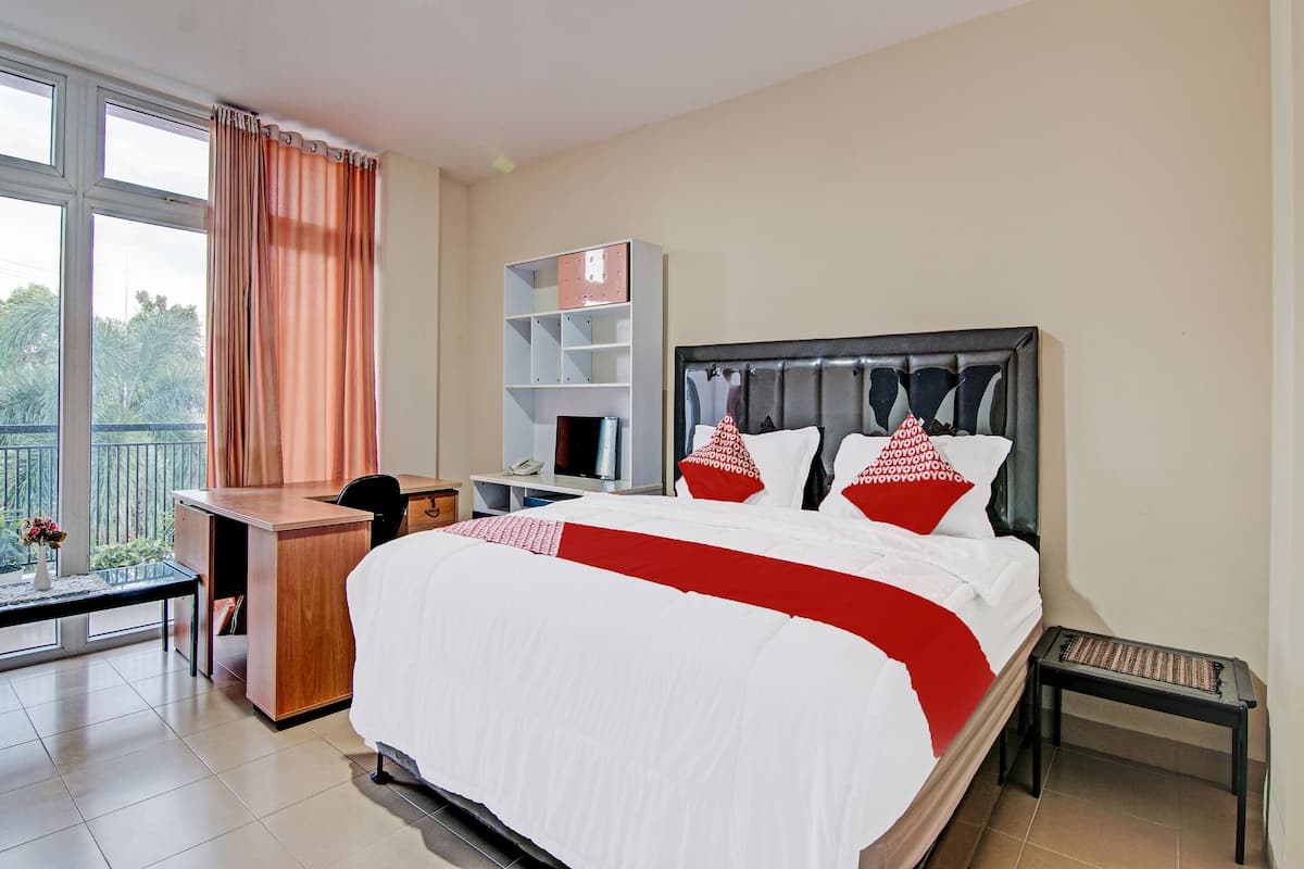 Habitación de hotel con una cama grande, un escritorio, una silla, una mesita, una estantería y un ventanal con cortinas.